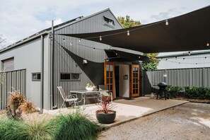 A modern outdoor patio area with a table and chairs, a barbecue grill, and a large shade sail. - Claret on Douro Three Cottages for Group Getaways (Mudgee)
