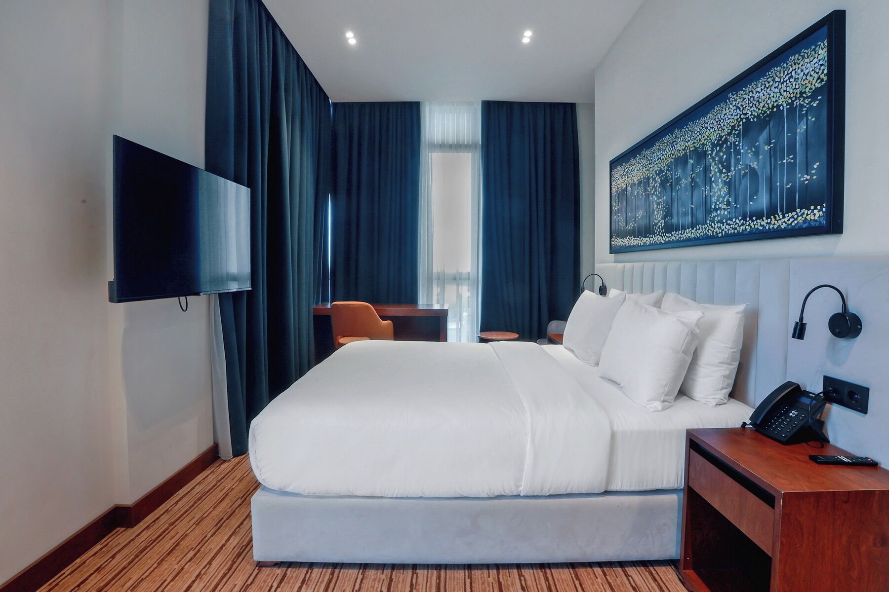 A hotel room with a bed, a desk, a chair, a TV, and a painting on the wall.
