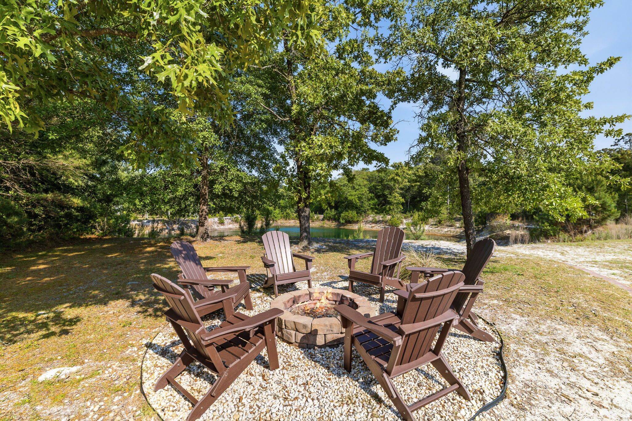 A fire pit with Adirondack chairs arranged around it, set on a gravel area with trees in the background.