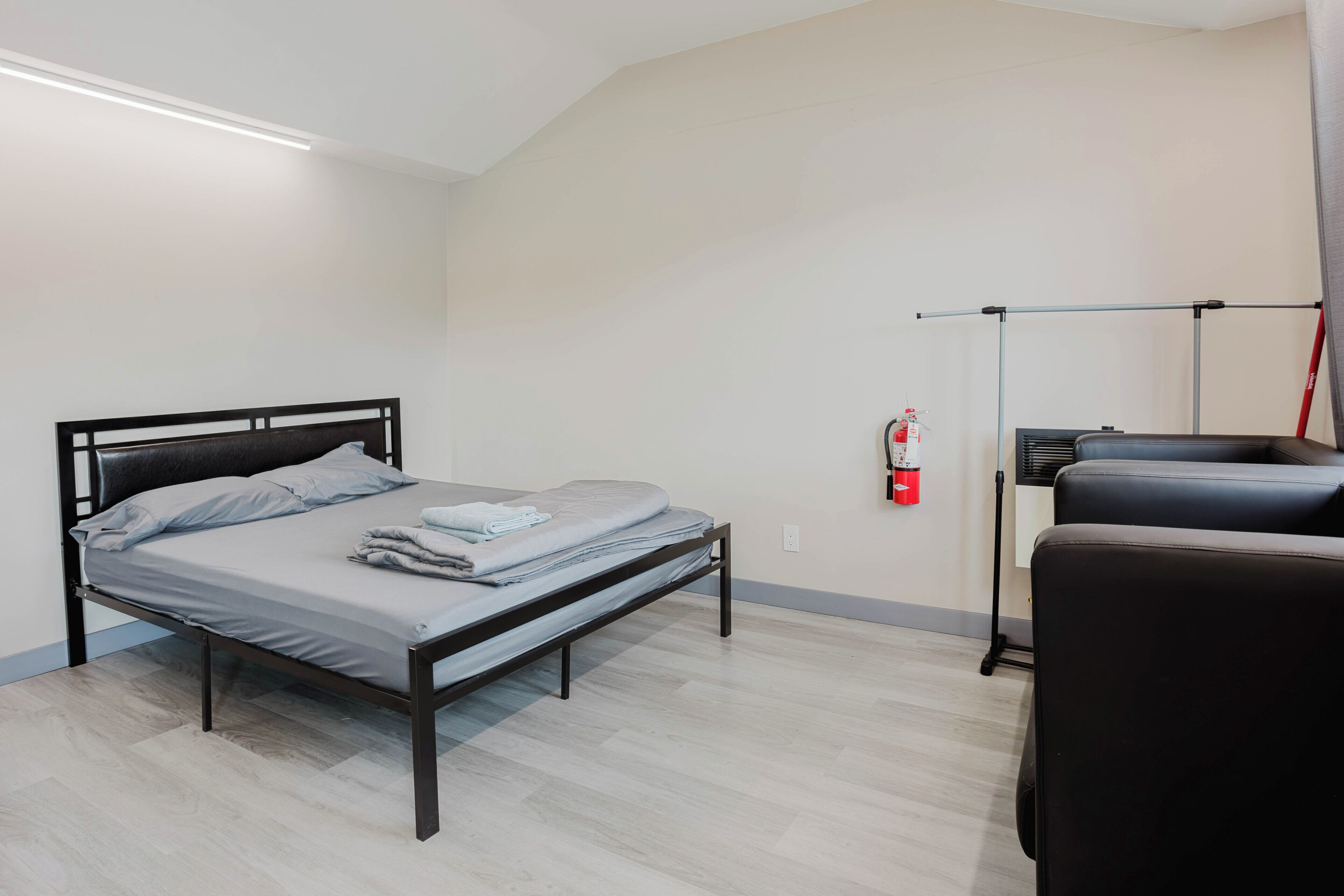 A single bed with a metal frame, a bedside table, a chair, a fire extinguisher, and a ceiling light fixture.