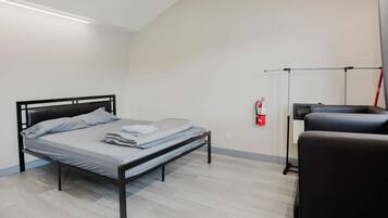 A single bed with a metal frame, a bedside table, a chair, a fire extinguisher, and a ceiling light fixture.