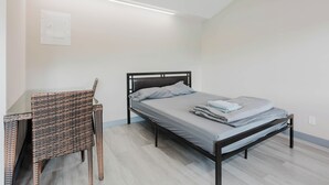 Un lit simple avec un cadre en métal, une petite table accompagnée d’une chaise en osier et un luminaire fixé au plafond.