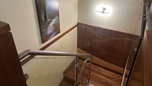 Uma escada de madeira com corrimão, um candeeiro fixado na parede e uma pintura representando uma cena de floresta.