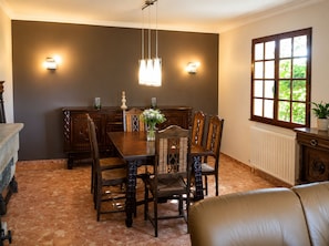 A dining room with a dark brown wall, a wooden dining table, six chairs, a chandelier, and a window with a view of greenery. - Domaine de la Barthe - Spacious Apartment in a Vineyard (Béziers)
