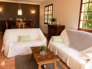 A living room with two white sofas, a wooden coffee table, a dining table with chairs, a radiator, and a window with greenery outside. - Domaine de la Barthe - Spacious Apartment in a Vineyard (Béziers)