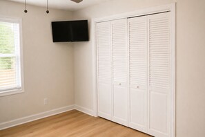 Una habitación con piso de madera, un armario blanco con puertas persianas y un televisor montado en la pared, además de una ventana con persianas.