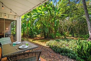 An outdoor dining area with a table and chairs, surrounded by lush greenery and banana plants. - Poipu Ground-Floor Gem | New A/C, Sleeps 6, Walk to Shopping & Dining (Koloa)