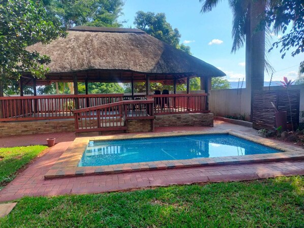 A pool area with a thatched-roof structure, a wooden deck, and a stone wall.