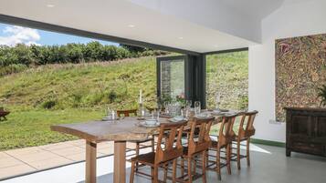 Un comedor con una mesa grande dispuesta para seis personas, sillas de madera y vistas a una colina cubierta de césped a través de un ventanal.