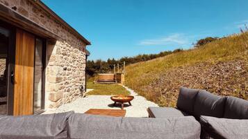 Un bâtiment en pierre avec une porte en bois, des sièges extérieurs dotés de coussins et une vue dégagée sur une colline.