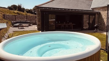 Un jacuzzi en un recinto de madera, un porche cubierto con mobiliario exterior y un edificio de piedra al fondo.