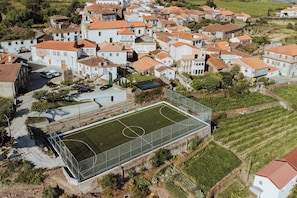 Luftaufnahme eines Wohngebiets mit weißen Gebäuden, einem eingezäunten Sportplatz und umgebender Bepflanzung.