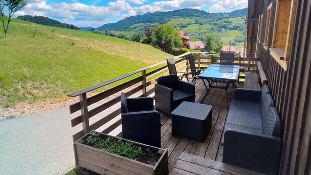 Une terrasse en bois avec des meubles d’extérieur, une vue sur un paysage verdoyant et un bâtiment en arrière-plan.