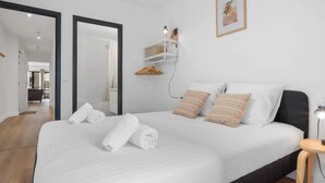 Uma cama bem arrumada com lençóis brancos, uma mesa de cabeceira, uma prateleira na parede com um abajur e um quadro emoldurado.