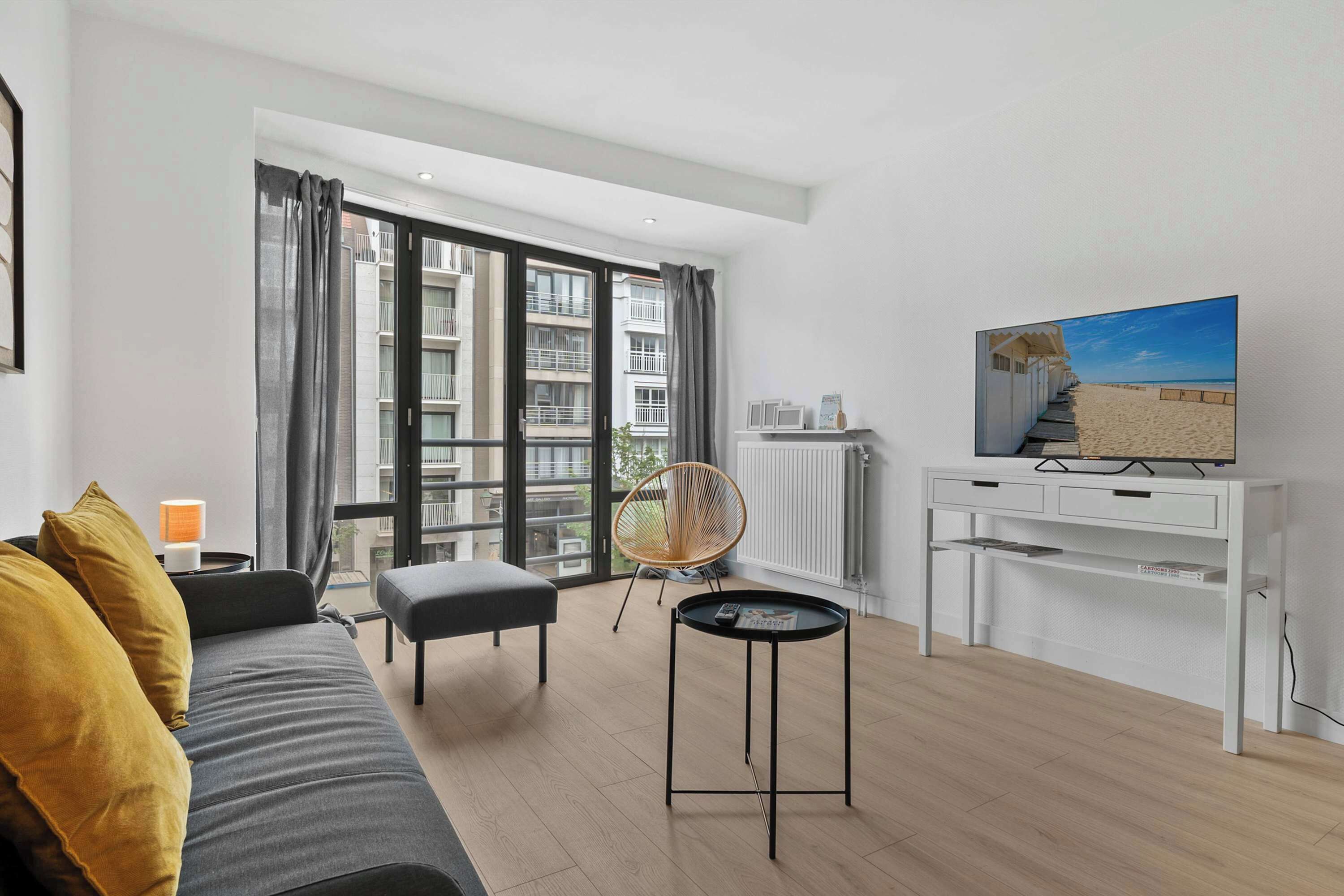 Un salon moderne avec un canapé, une petite table, un fauteuil, un téléviseur et une vue sur un immeuble à travers de grandes fenêtres.