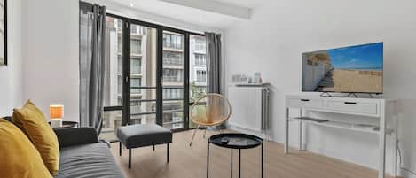 A modern living room with a sofa, a small table, a chair, a TV, and a view of a building through large windows.