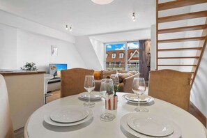 A dining area with a round table set for four, a sofa in the background, and a view of a residential building through large windows. - Casiola - Charming Duplex with Terrace in Knokke (Knokke-Heist)