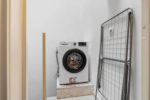 Eine kompakte, weiße Waschmaschine auf einer kleinen Plattform, eine Maschendrahttür und eine schlichte Wand.