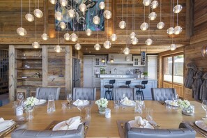 Une salle à manger spacieuse avec une grande table en bois dressée pour six personnes, une cuisine en arrière-plan et de magnifiques lustres en cristal suspendus au plafond.