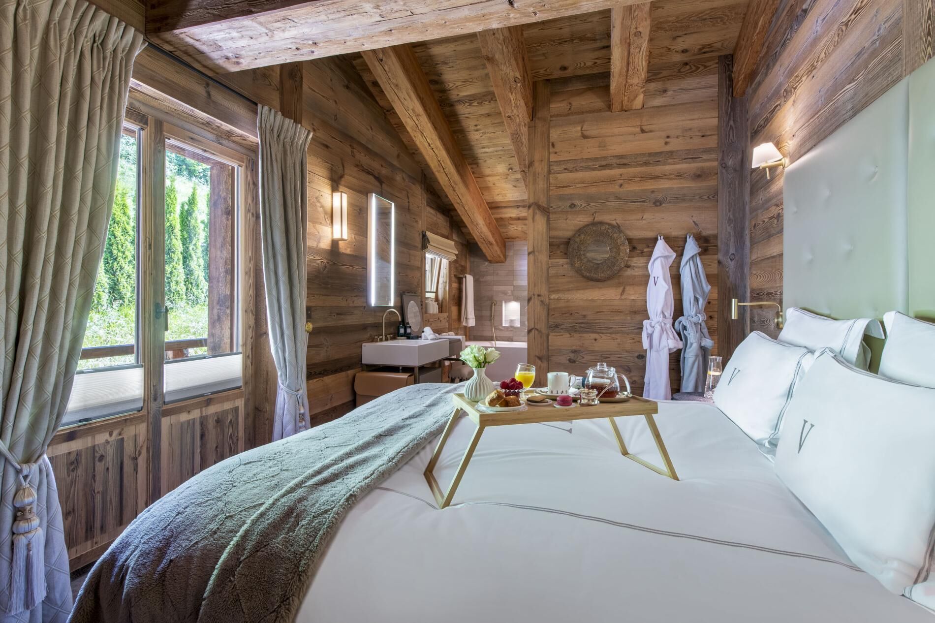 Une chambre dans un chalet en bois, avec un lit, un plateau de petit-déjeuner, un lavabo et une fenêtre avec des rideaux.