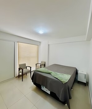 Ein Schlafzimmer mit einem Bett, einem Sessel, einem Schreibtisch und einem Fenster mit Jalousien.
