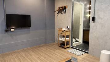 A modern bathroom with a wooden floor, a mounted TV, a towel rack, a shower area, and a small table with a mat.