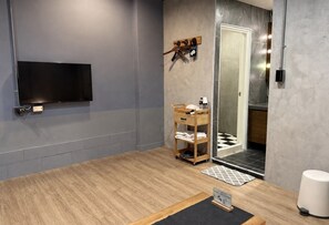A modern bathroom with a wooden floor, a mounted TV, a towel rack, a shower area, and a small table with a mat.
