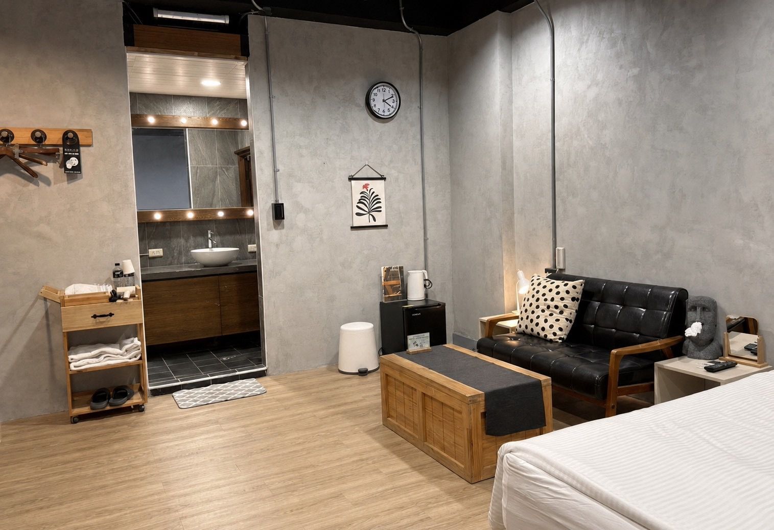 A modern room with a bed, a sofa, a small table, a wooden cabinet, a sink, and a clock on the wall.