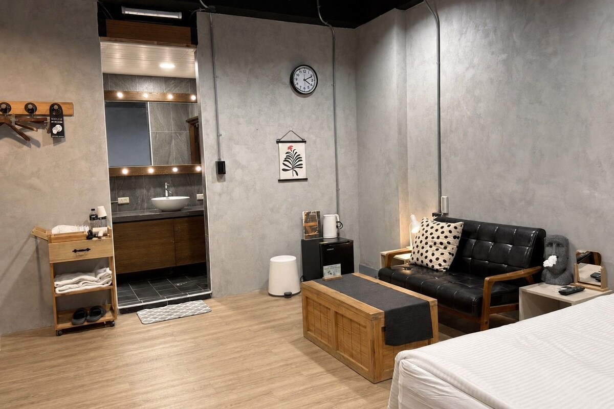 Ein modernes Zimmer mit Bett, Sofa, kleinem Tisch, Holzschrank, Waschbecken und einer Wanduhr.