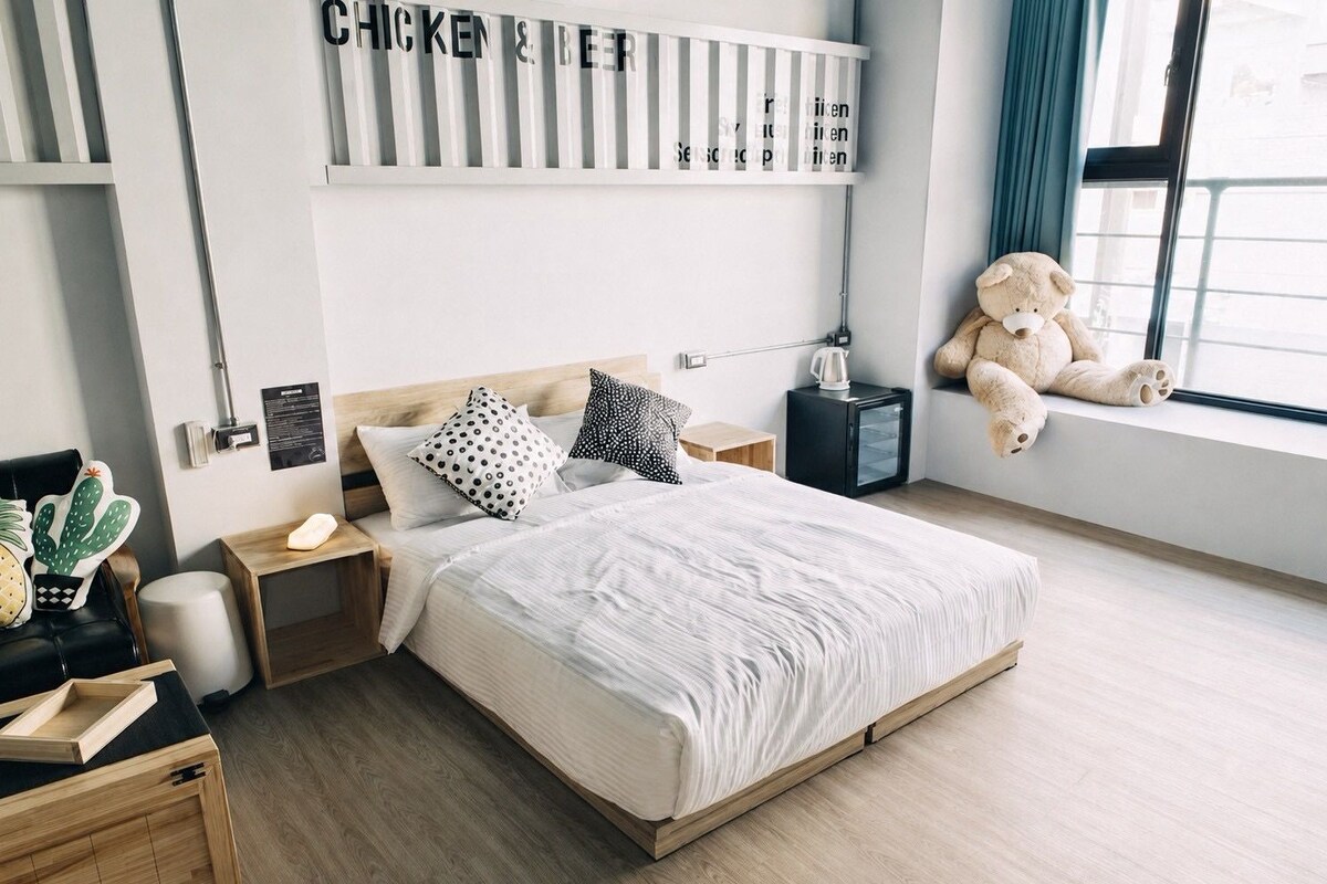 Ein modernes Schlafzimmer mit einem Bett, Nachttischen, einem Wandregal, einem Fenster mit Teddybär und Zimmerpflanze.