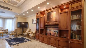 A living room with a large wooden entertainment center, a flat-screen TV, and a bookshelf. - New Cairo,vast classy 3 BR Apt.in quite villa inside fruity garden. Guarded 24/7 (New Cairo City)