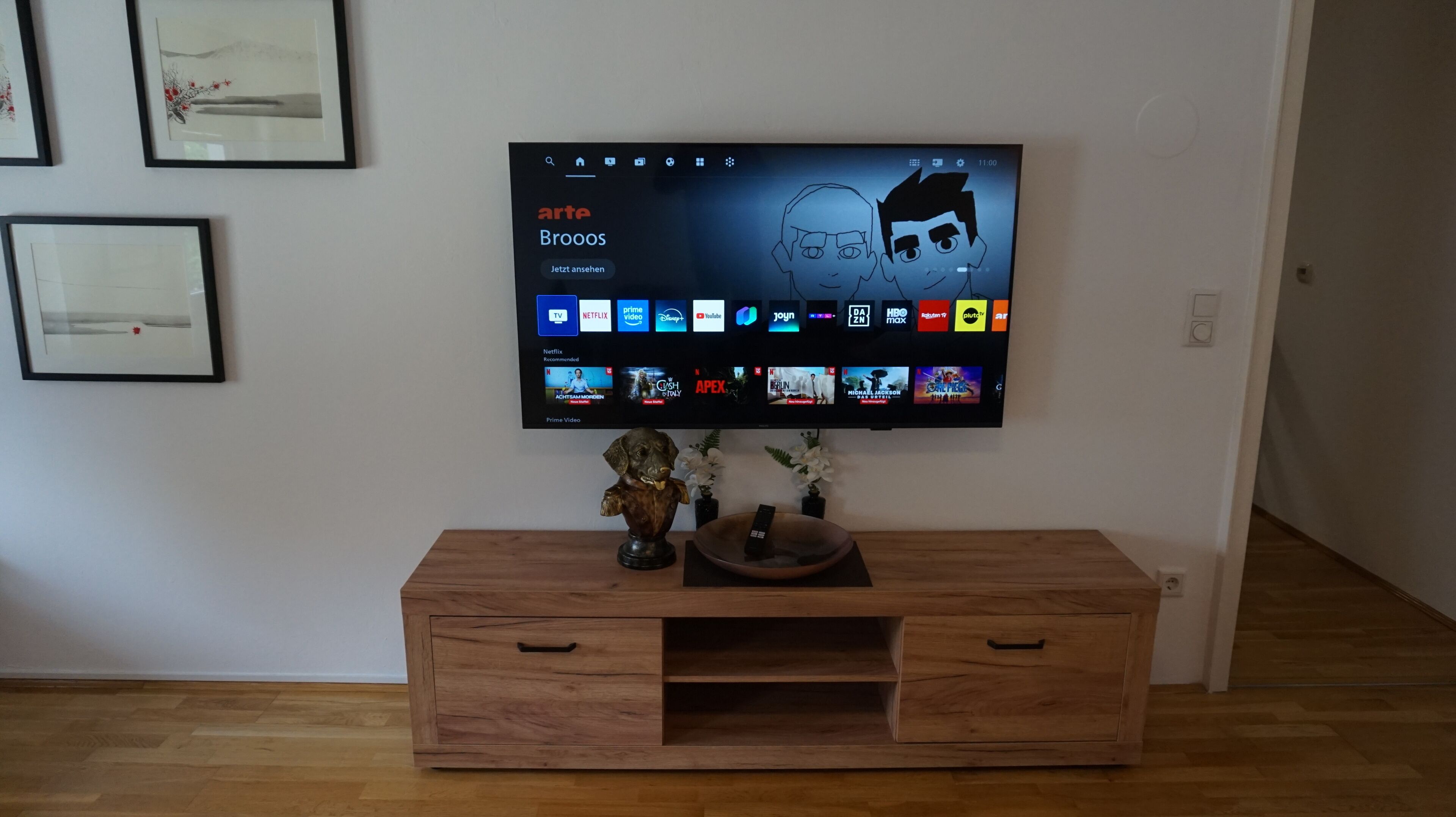 Ein Flachbildfernseher an der Wand über einem Holz-TV-Schrank mit Fernbedienung und Dekorationselementen.