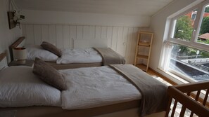 Ein Schlafzimmer mit zwei Betten, einem Nachttisch mit Lampe, einer Holzablage und einem Fenster mit Blick auf Gebäude.