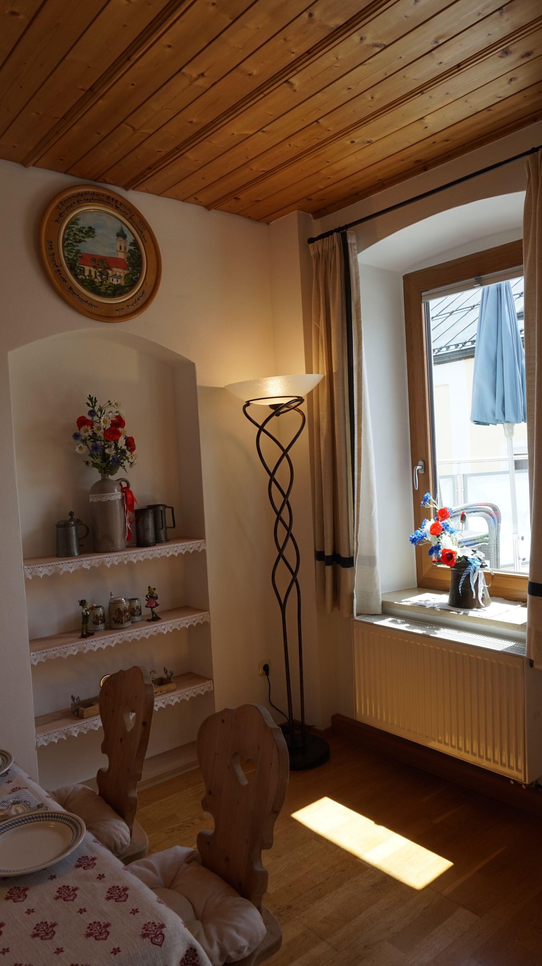 Ein gemütliches Zimmer mit Holzboden, einer Stehlampe, einem Fenster mit Vorhängen, einem Regal mit Dekoration und einem Sessel mit Tisch.