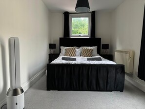 A bedroom with a large bed, two bedside tables, a window, a radiator, and a standing heater. - Entire Apartment king size bed near Town Centre (Colchester)