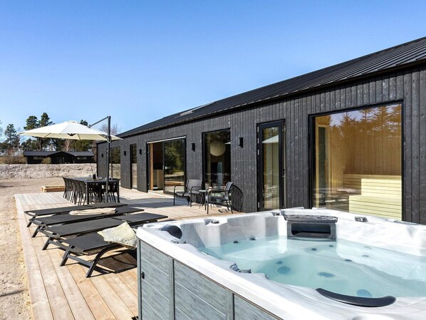 A modern outdoor hot tub area with lounge chairs and a table, adjacent to a building with large glass doors. - 5 Star Holiday Home in Saeby (Sæby)