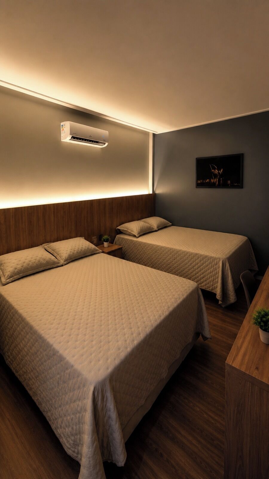 A hotel room with two beds, a bedside table with a plant, a wall-mounted air conditioner, and a framed picture on the wall.