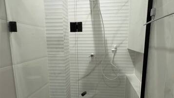 A glass-enclosed shower stall with a black frame, white tiled walls, and a white toilet to the left.