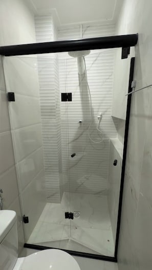 Cabina de ducha con paredes de azulejos blancos, cerramientos de cristal y un inodoro blanco a la izquierda.