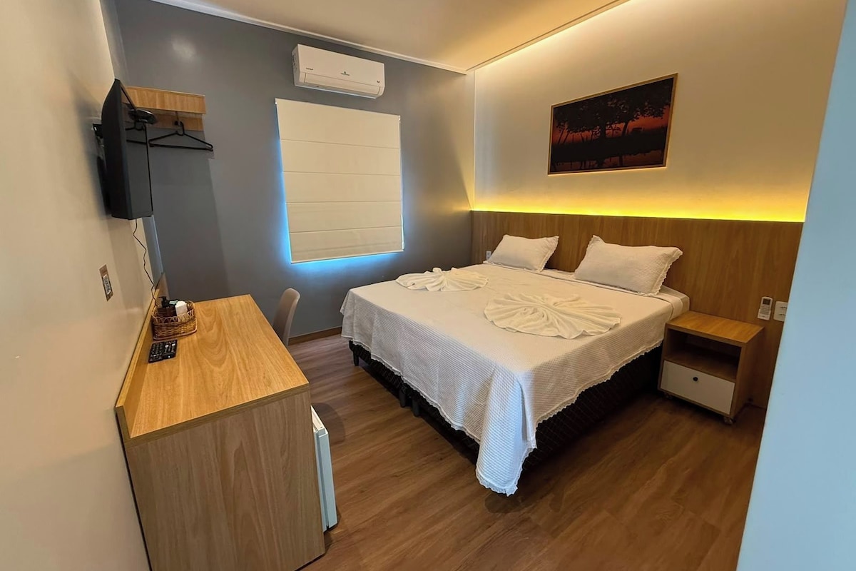 A hotel room with a bed, bedside table, a desk with a chair, a television, and a window with blinds.
