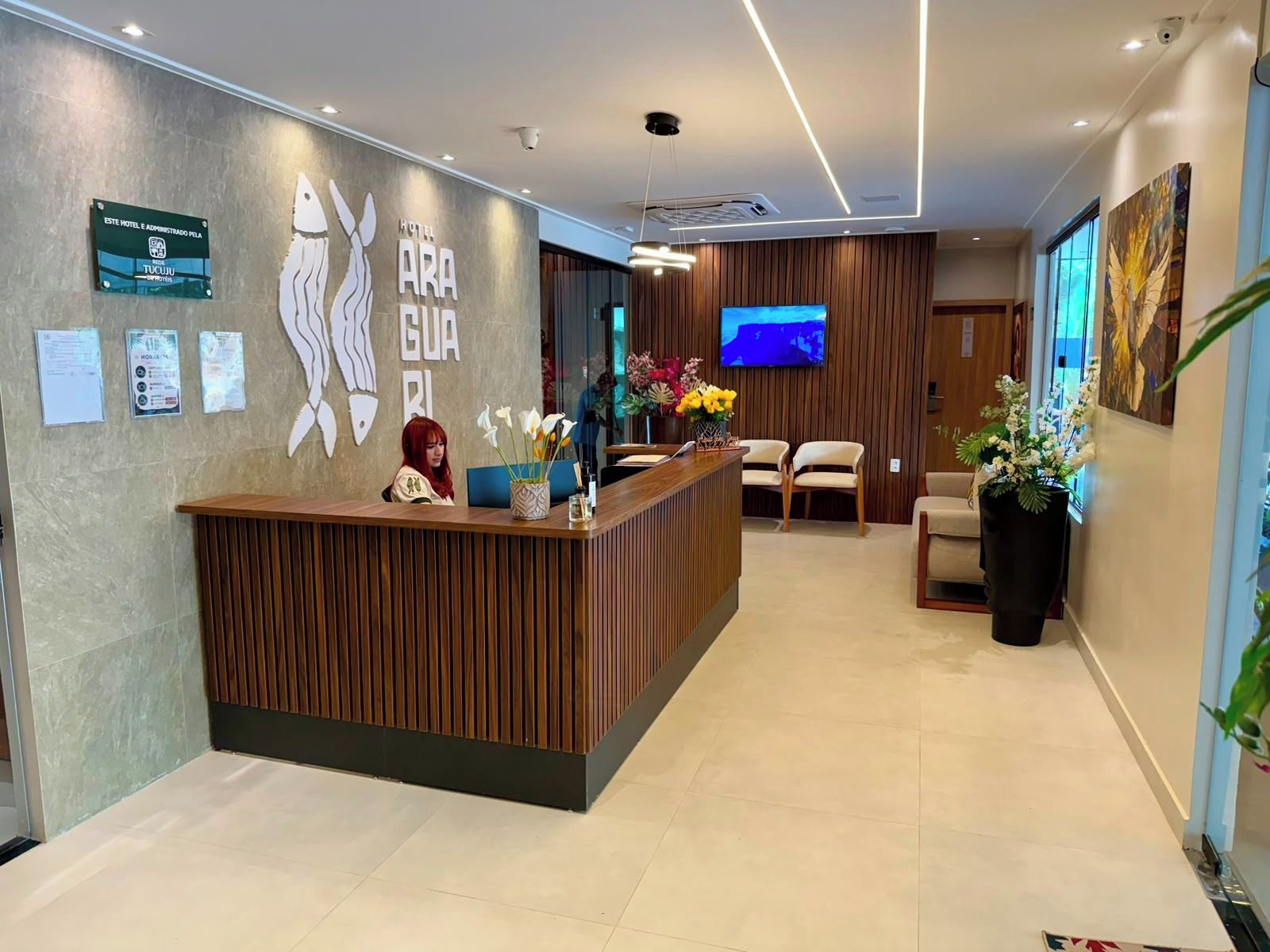 Een hotel receptie met een houten balie, een receptionist, een flatscreen televisie en een grote potplant.
