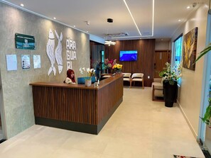 A hotel reception area with a wooden reception desk, a receptionist, a flat-screen TV, and a large potted plant. - Araguari Hotel Ferreira Gomes  (Ferreira Gomes)