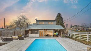 Uma piscina retangular com superfície de água azul cristalina, rodeada por um terraço pavimentado com mobiliário de exterior e uma área de estar coberta.