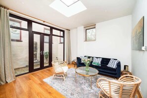 A modern living room with a dark sofa, wicker chairs, a glass coffee table, and a large sliding glass door leading to an outdoor area. - Unique 2 Bed Chelsea townhouse | BeeMyGuest® (London)