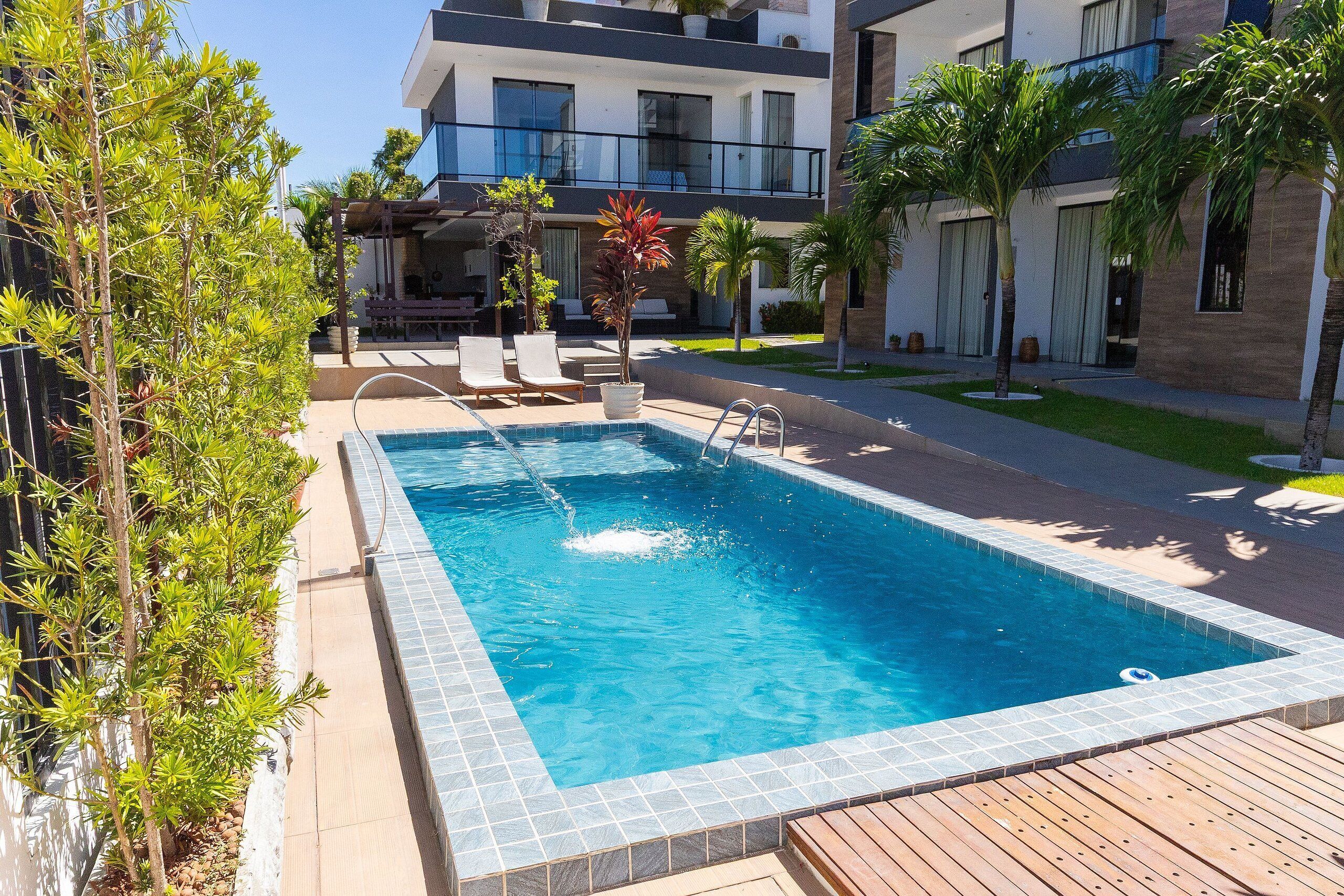 Une piscine dotée d’une terrasse en bois, entourée de plantes en pot et bordée d’un bâtiment moderne avec des balcons.