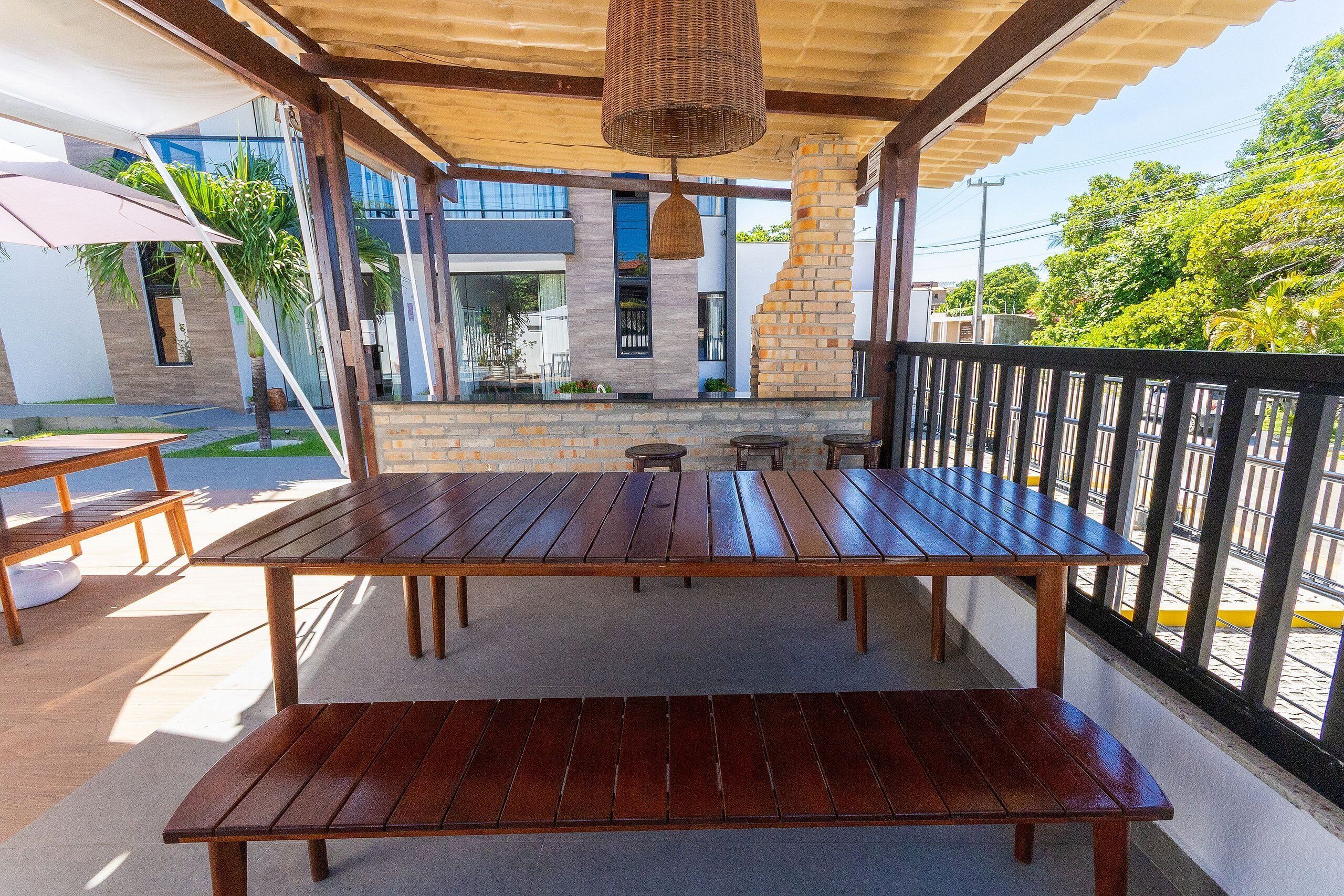 A covered outdoor seating area with a wooden table and benches, a hanging light fixture, and a view of a modern building with a stone wall and glass windows.