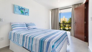 A bedroom with a bed, a painting, a window with curtains, and a view of buildings. - Altos do Caju - Triplex in Pirangi 300 Meters From the World's Largest Cashew Tree by Carpediem (Parnamirim)