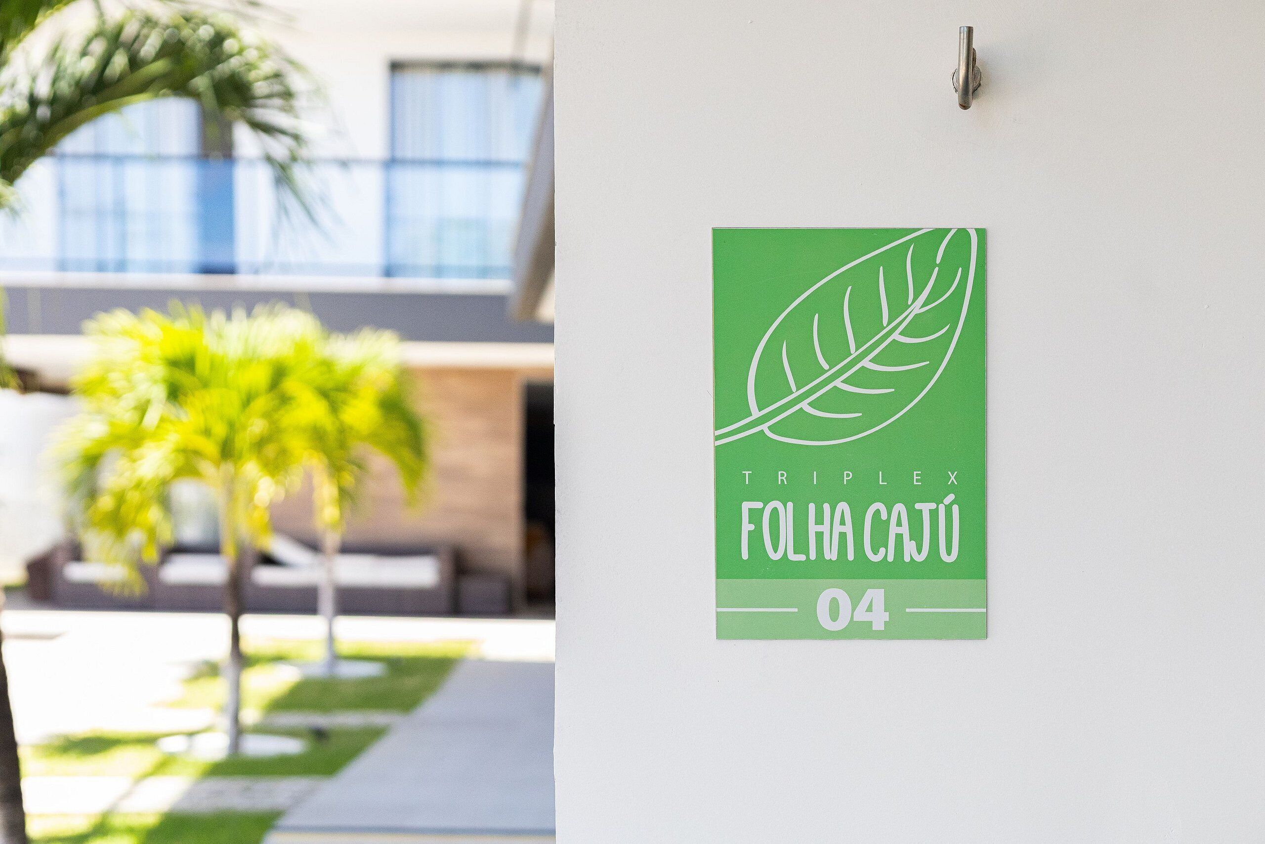 Un letrero verde con una hoja y texto sobre una pared blanca, una palmera y un edificio con ventanas al fondo.