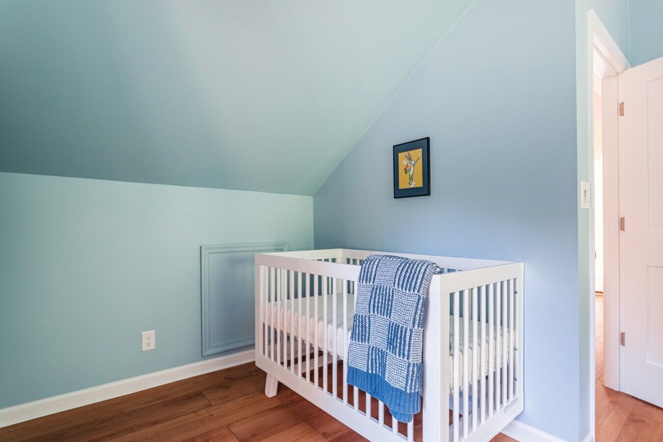 A room with a white crib, light blue walls, a framed picture, and a door.