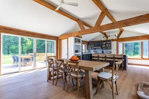 Amplio interior de madera con comedor, cocina y grandes puertas corredizas de vidrio que dan a una terraza exterior.